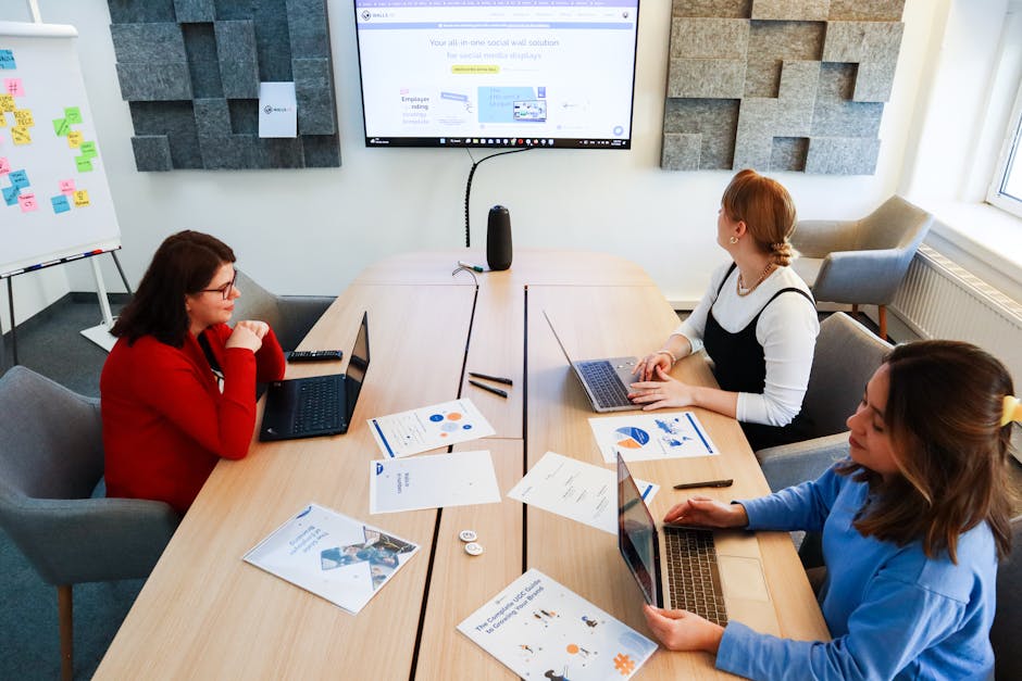 Three women collaborating in a modern office with laptops and presentation materials.